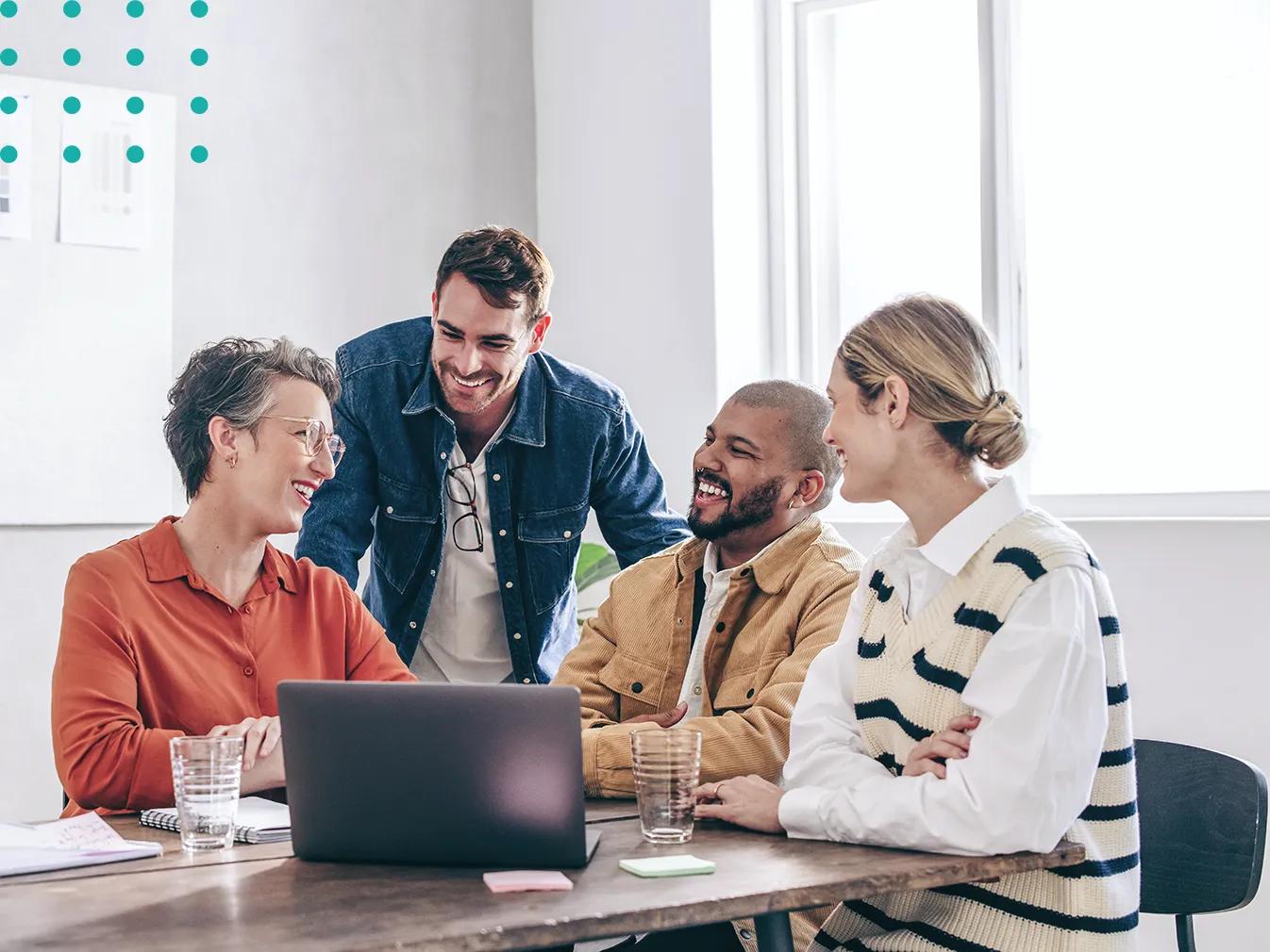 people laughing while crowding around a laptop on a conference table